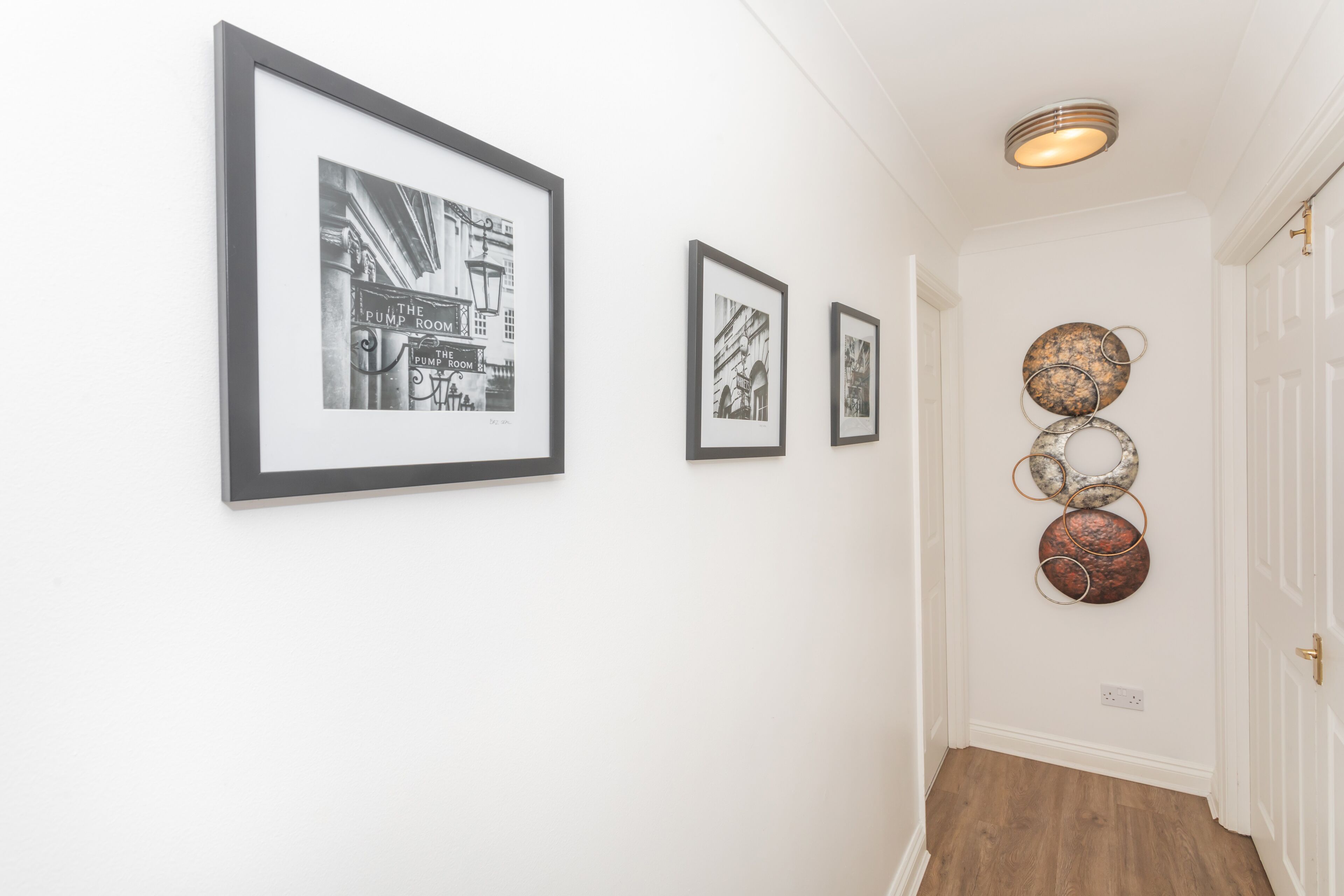 A hallway with framed black and white photographs on the wall, a decorative metal piece, and a door at the end.