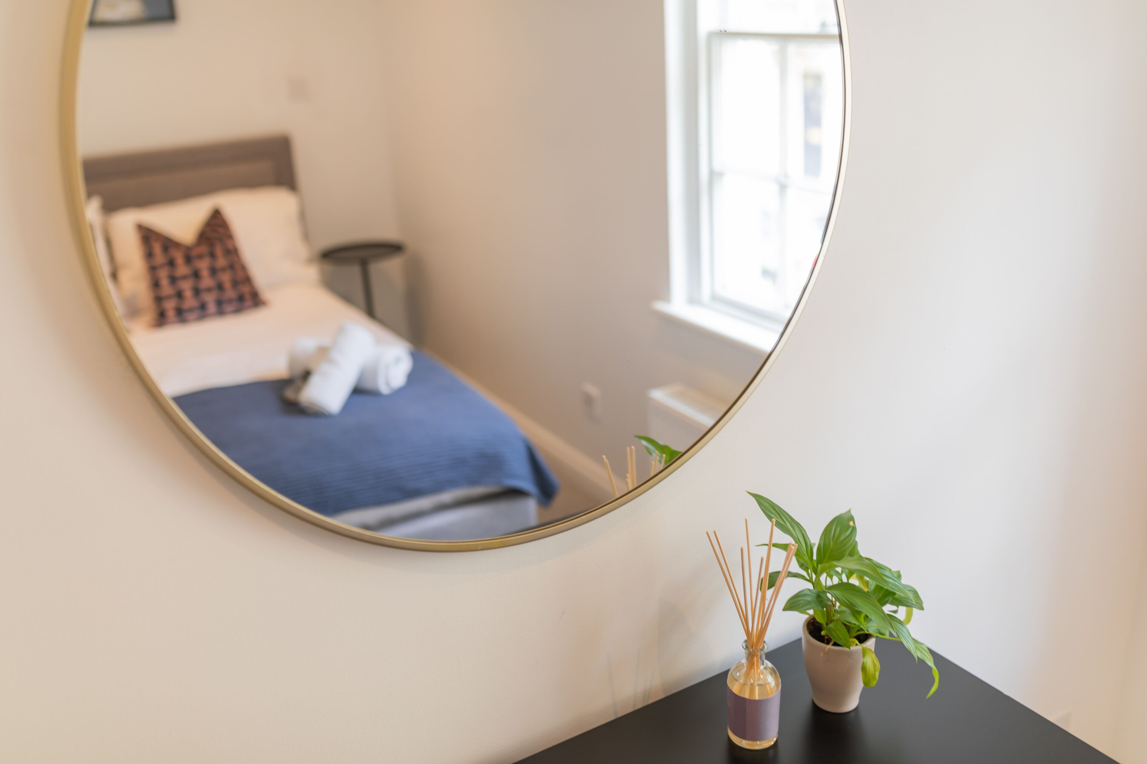 A bedroom with a bed, a bedside table with a potted plant, and a mirror reflecting the room.
