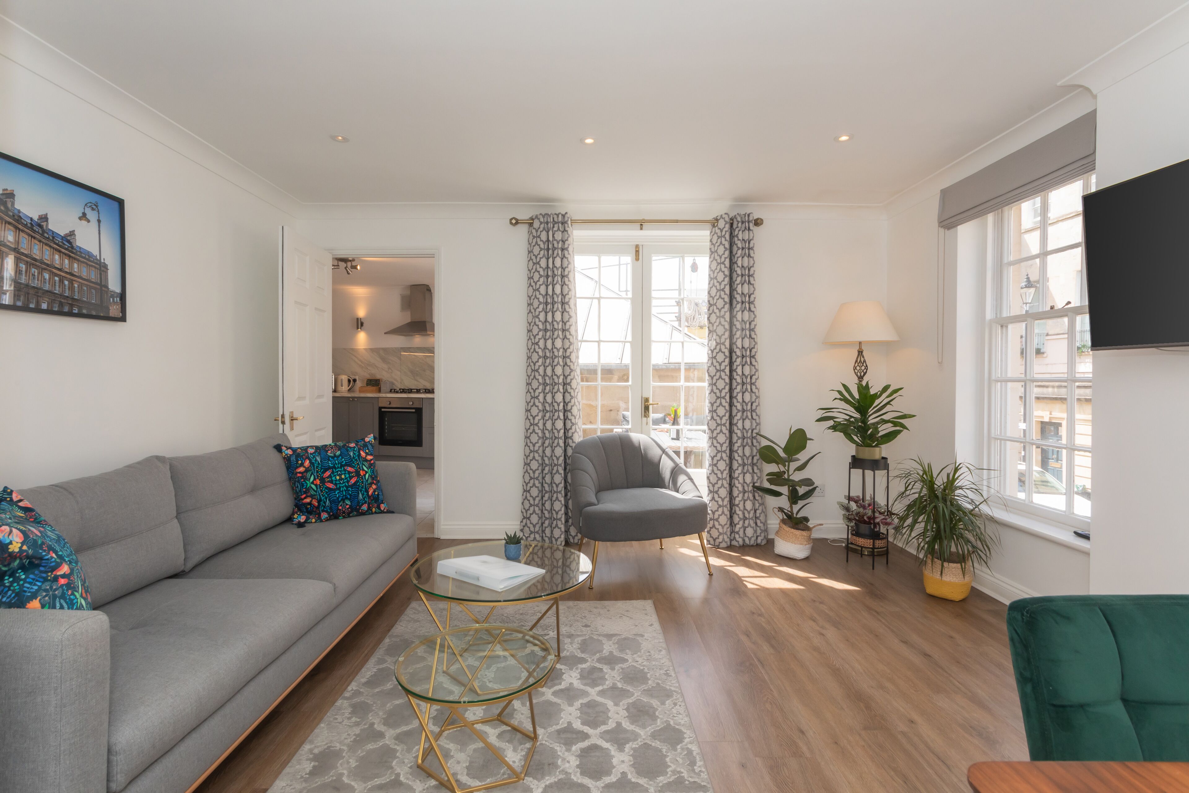 A modern living room with a grey sofa, a glass coffee table, a grey armchair, and a green armchair. There is a TV mounted on the wall, a painting of a building, and a window with curtains.