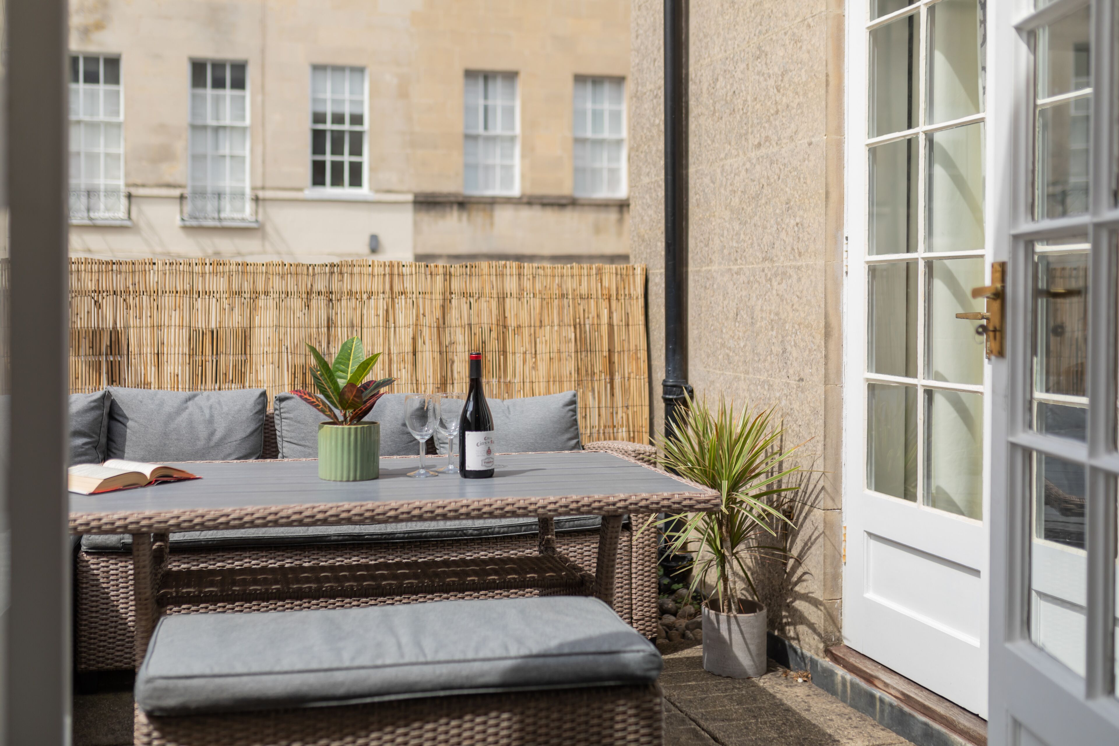 A small outdoor seating area with a table, cushions, and a bottle of wine.