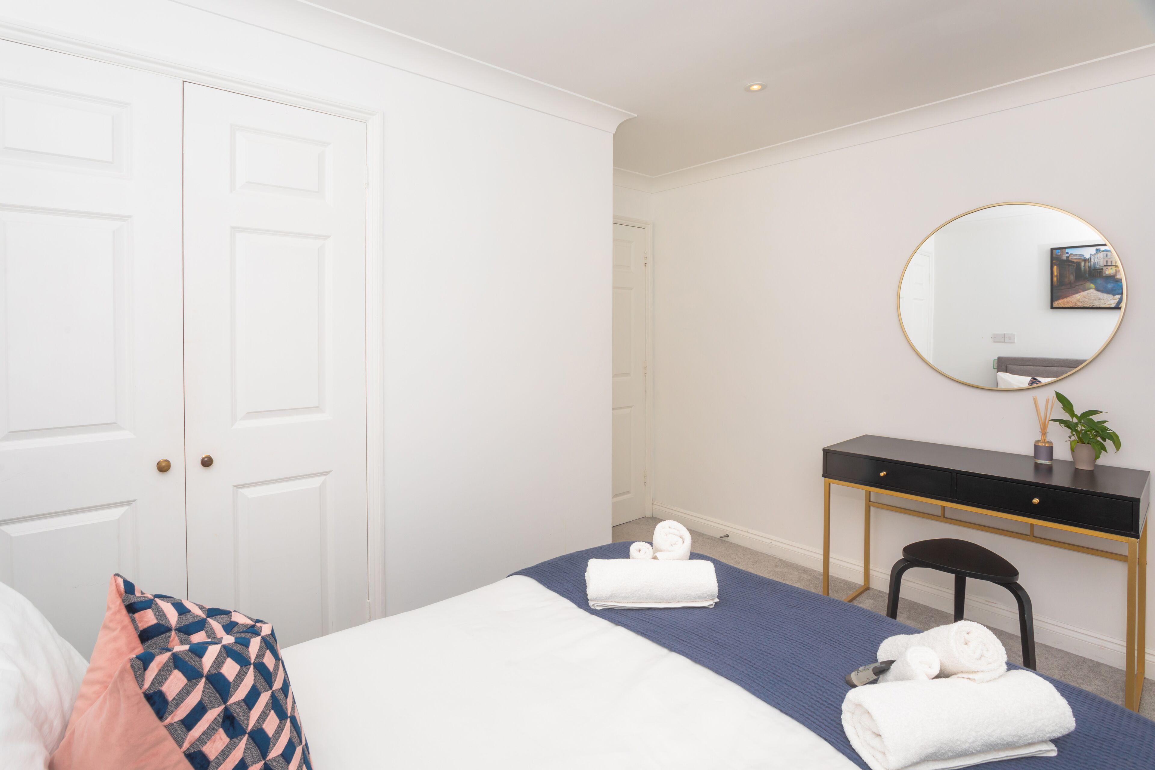 A hotel room with a bed, a desk, a chair, a round mirror, and a door.