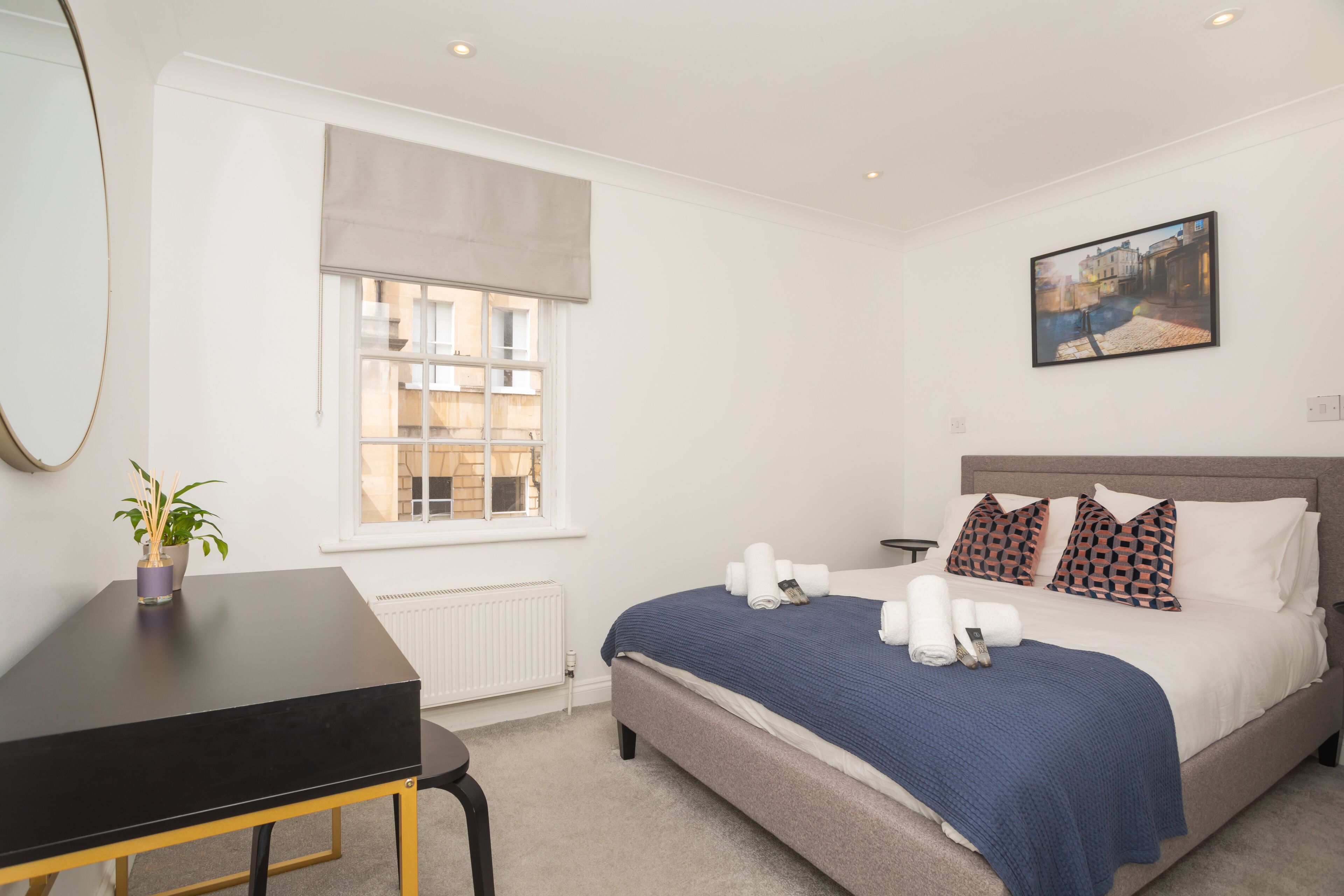 A bedroom with a bed, a bedside table, a window with a view of buildings, and a painting on the wall.