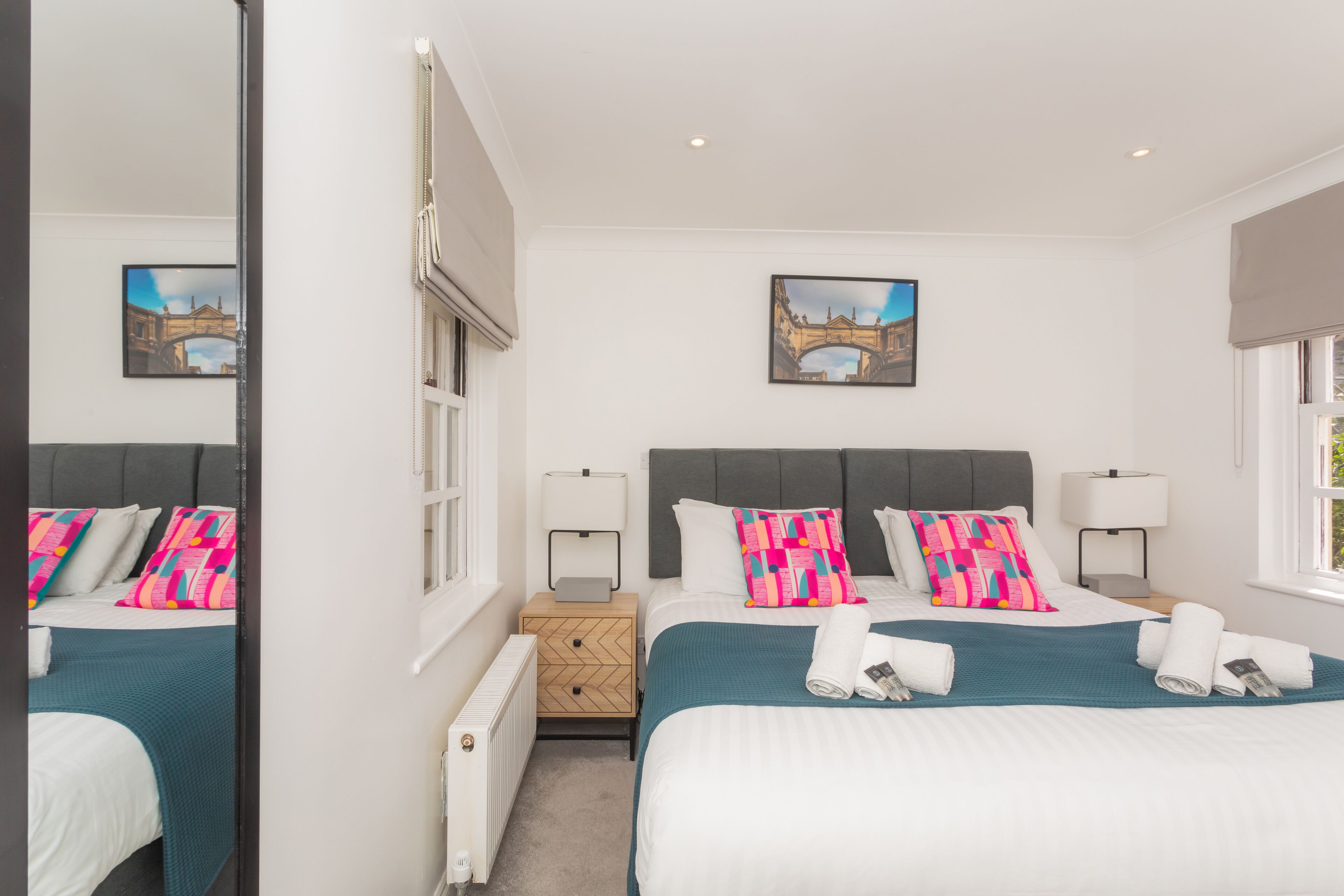 A double bed with teal bedding, two bedside tables with lamps, a mirror, and a framed picture of a bridge on the wall.