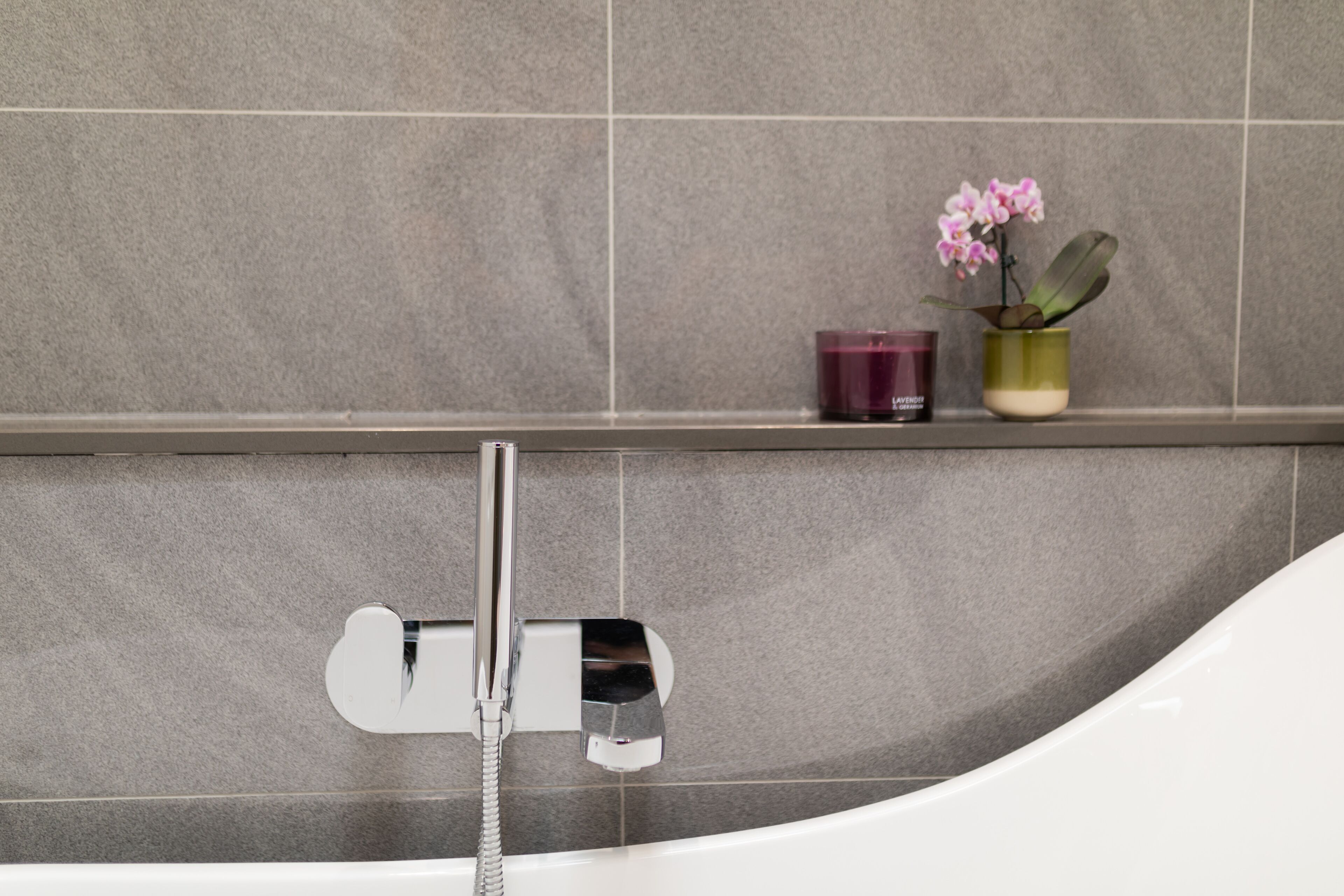 A bathtub with a chrome faucet and a handheld showerhead, a shelf with a purple candle and a potted orchid, and a tiled wall.