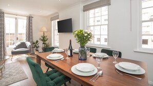 Un comedor con una mesa de madera dispuesta para cuatro personas, rodeada de sillas tapizadas de verde. En la mesa hay un jarrón con flores y se ve una botella de vino.