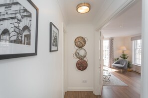 A hallway with a framed black and white photo, a wall-mounted circular metal art piece, a grey armchair, and a potted plant. - Crescent Mews (Bath)