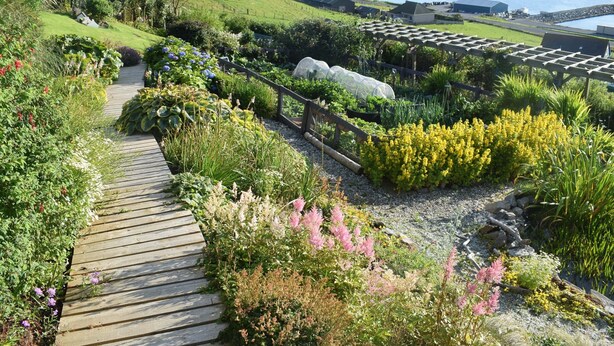 Un sendero en un jardín conduce a un área de embarcadero con barcos, rodeado de exuberante vegetación y vistas al agua.
