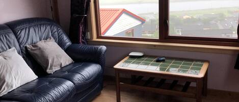 Una sala de estar con un sofá de cuero, un tapete morado, una mesa de centro de madera con un juego de mesa y una ventana con vistas a una casa y al agua.