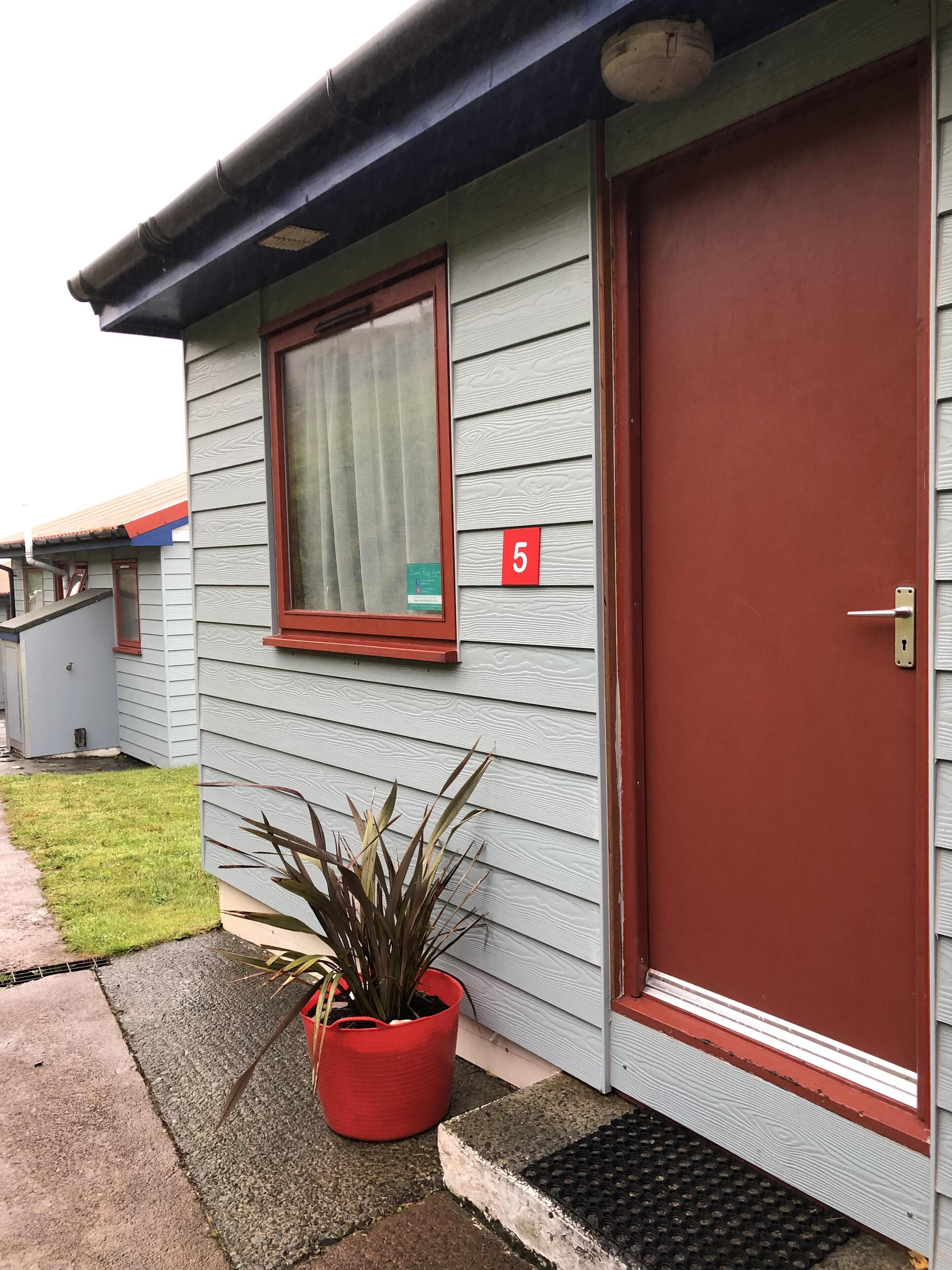 A single-story building with a red door and a window with a curtain. A red number '5' is visible on the wall.