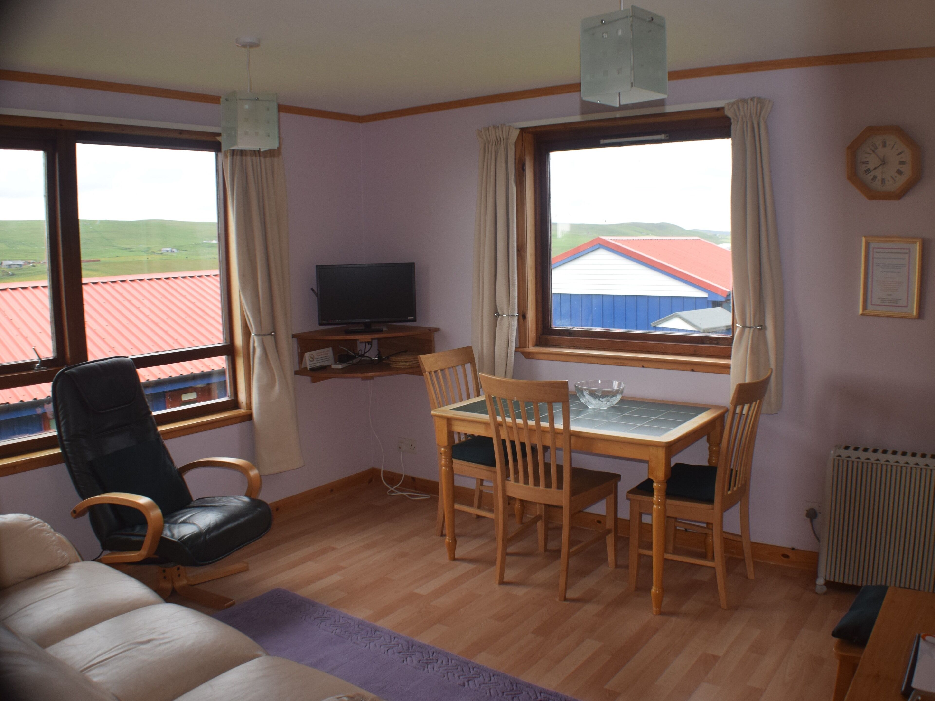 A room with a wooden dining table, chairs, a television on a shelf, a sofa, and large windows with curtains.