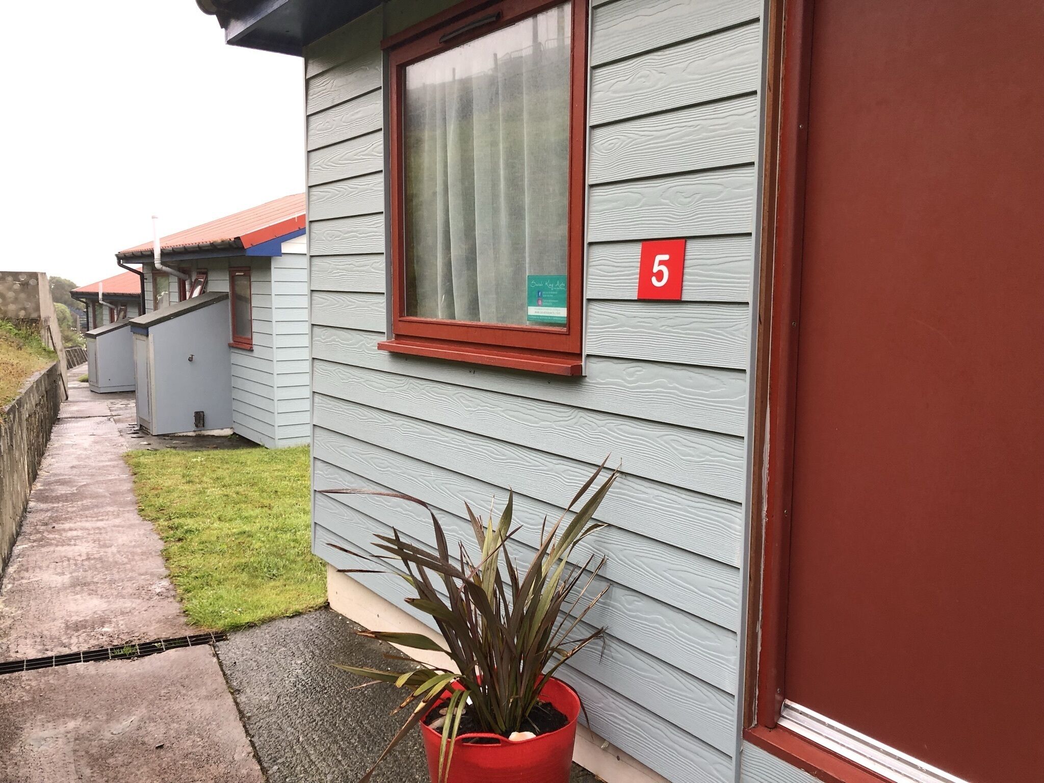 Fila de cabañas de un piso con un cartel con el número 5, una pequeña ventana con cortina y una planta en maceta al frente.