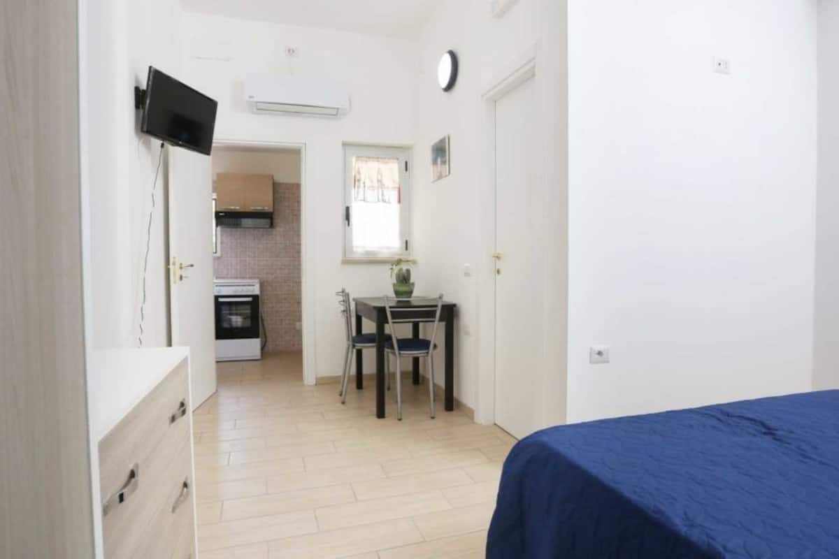 A compact room with a bed, a desk, and a chair. There is a kitchenette in the background.