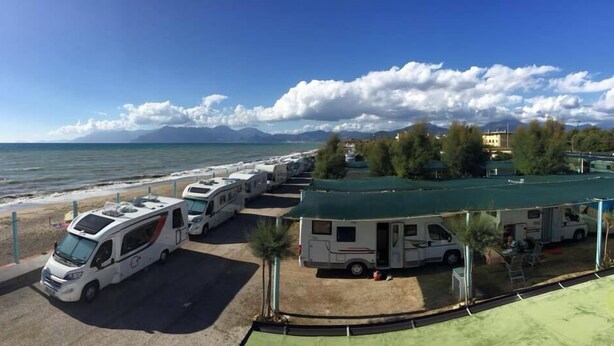 Una hilera de vehículos recreativos estacionados junto a la playa, con vistas despejadas al mar y a las montañas al fondo.