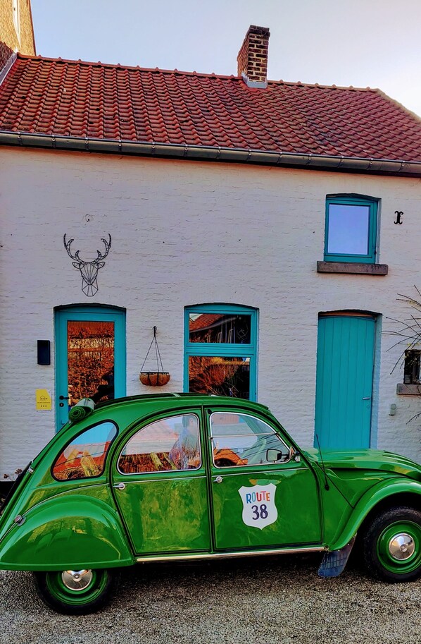 A vintage green car parked in front of a building with a red-tiled roof, a white wall, and a blue door. - Wetterdelle (Heers)