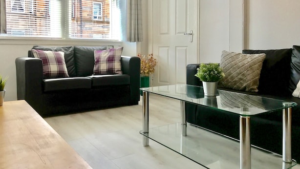 A modern living room with a glass coffee table, a black leather sofa with plaid cushions, and a view of a building through the window blinds. - Easter Road Edinburgh Apartment (Edinburgh)
