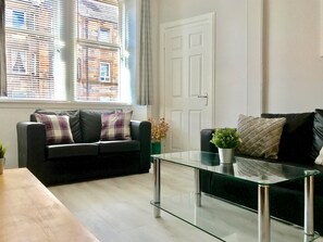 A modern living room with a glass coffee table, a black leather sofa with plaid cushions, and a view of a building through the window blinds. - Easter Road Edinburgh Apartment (Edinburgh)