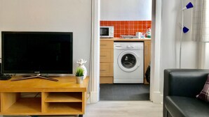 A modern living room with a flat-screen TV, wooden entertainment unit, and a small plant. A black sofa and a black leather couch are visible. The room leads to a kitchen with a washing machine, microwave, and a clock on the wall. - Easter Road Edinburgh Apartment (Edinburgh)