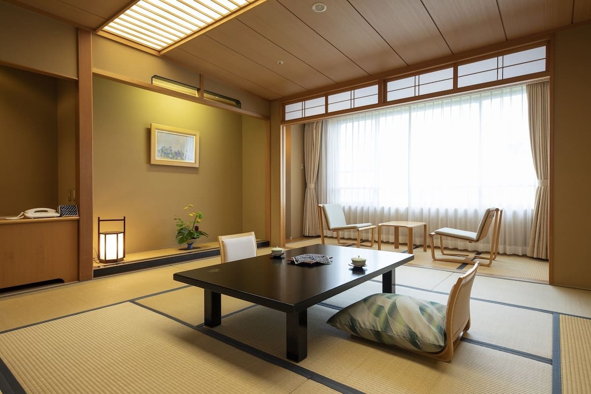 A traditional Japanese-style room with tatami flooring, a low table, chairs, and a large window with curtains.