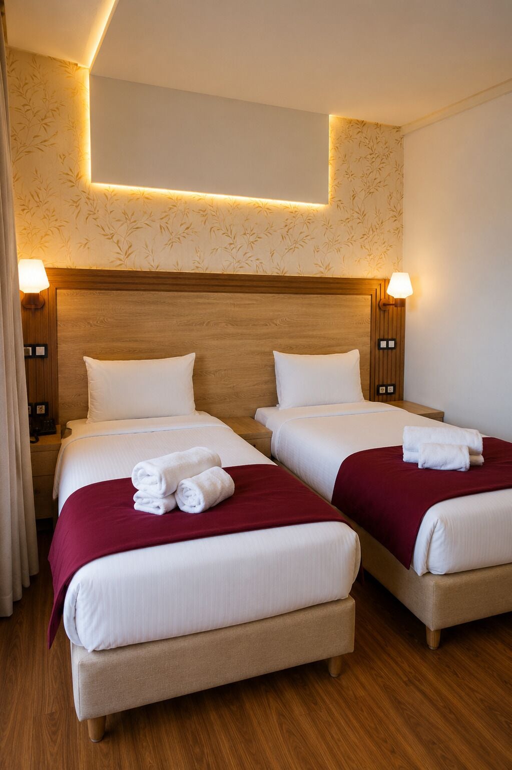 A hotel room with two beds, each with white linens and red blankets, wooden headboards, and wall-mounted lamps.