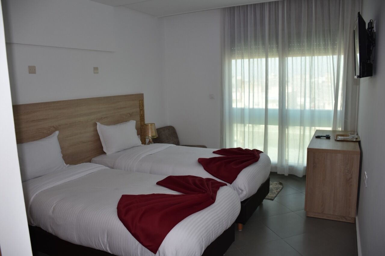 A hotel room with two beds, a wooden headboard, a bedside table with a lamp, a TV mounted on the wall, and a large window with sheer curtains.