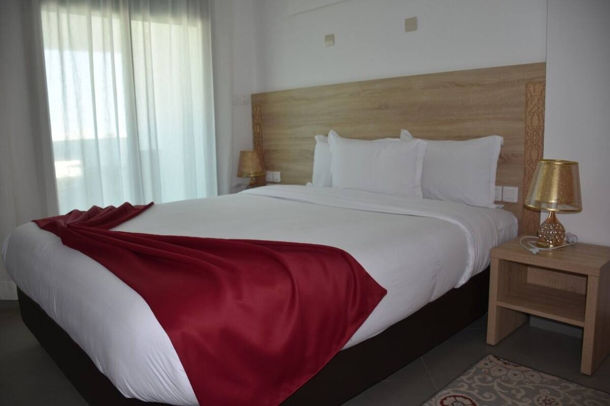A hotel room with a large bed, a wooden headboard, two bedside tables with lamps, and a window with sheer curtains.