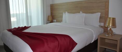 Una habitación de hotel con una cama grande, un cabecero de madera, dos mesitas de noche con lámparas y una ventana con cortinas translúcidas.