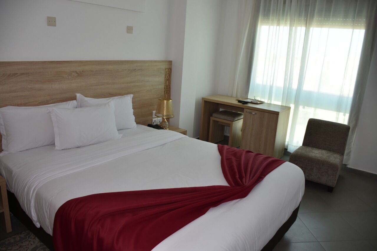 A hotel room with a large bed, a wooden headboard, a desk with a lamp, a chair, and a window with curtains.
