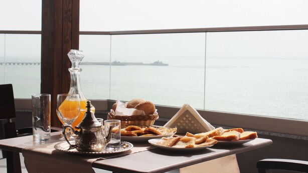 A table with a glass of orange juice, a bottle, a basket of bread, and a plate of pastries, with a view of the sea and a ship in the distance. - Dakhla Sur Mer (Dakhla)