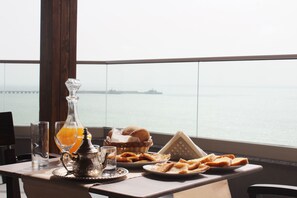 Uma mesa com um copo de sumo de laranja, uma garrafa, uma cesta de pão e um prato de pastelaria, com vista para o mar e um navio ao longe.
