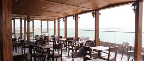 Un espacio de comedor al aire libre cubierto con mesas y sillas de madera, amplias ventanas que ofrecen vista al mar y un puente distante.