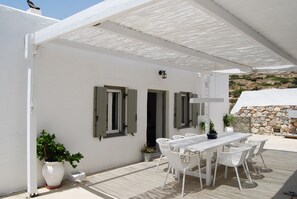 A covered outdoor patio with a white table and chairs, a potted plant, and a stone wall in the background. - Maistrali Studio - House Levante (Syros)