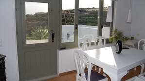 A dining area with a white table and chairs, a view of the countryside through large windows, and a door with a view. - Maistrali Studio - House Levante (Syros)