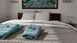 Een hotelkamer met een groot bed, witte lakens, een tropisch schilderij boven het bed en twee opgevouwen handdoeken op het bed.