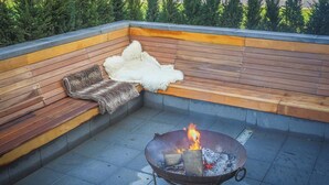 Een terras op het dak met vuurkorf, houten bank en een bont plaid.