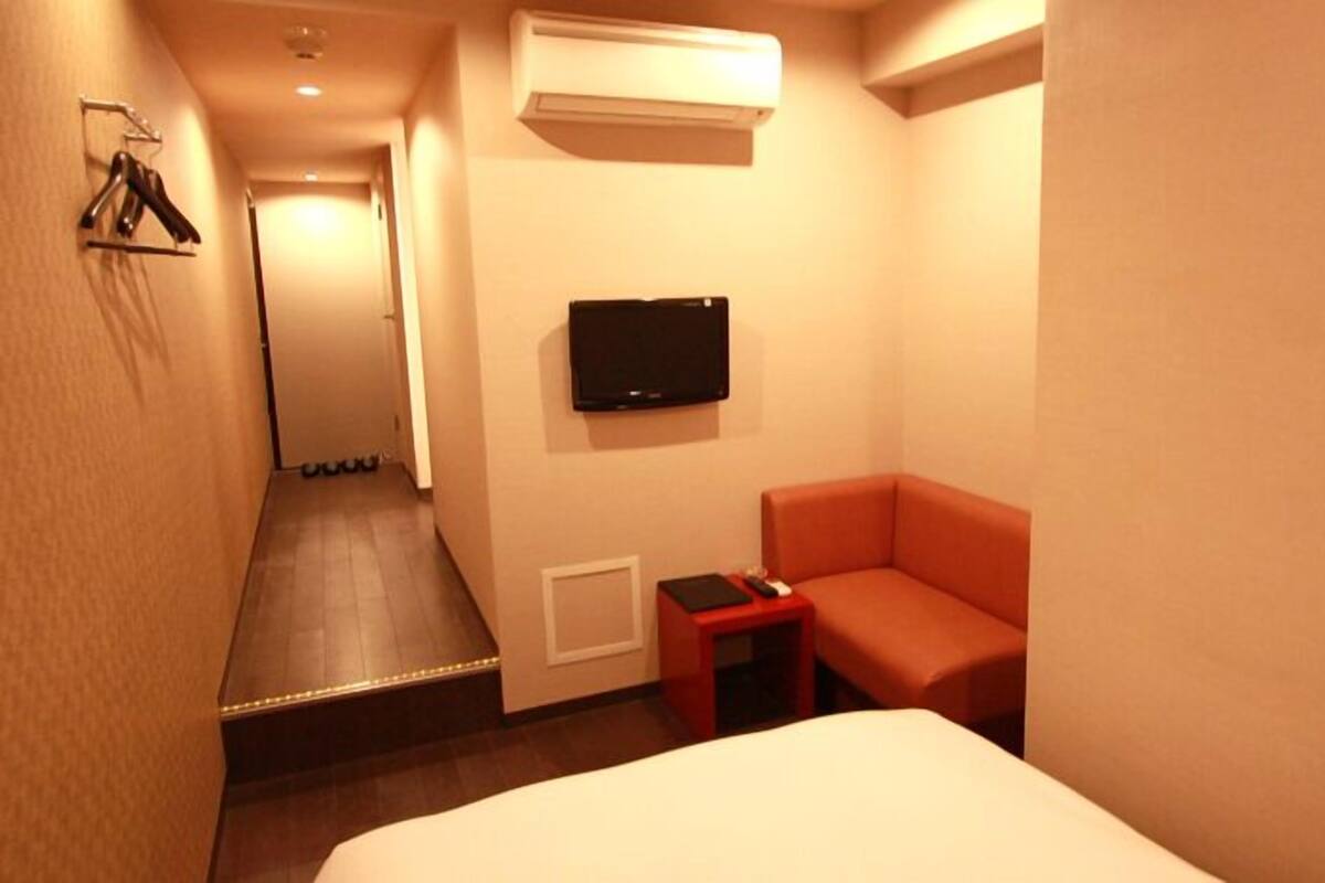 Habitación de hotel con una cama, un sillón rojo, un televisor montado en la pared y una mesita con un control remoto.