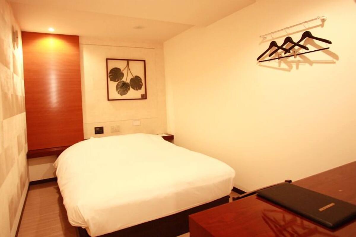 Habitación de hotel pequeña y ordenada con una cama individual, una mesita de noche, perchero en la pared y un cuadro enmarco.