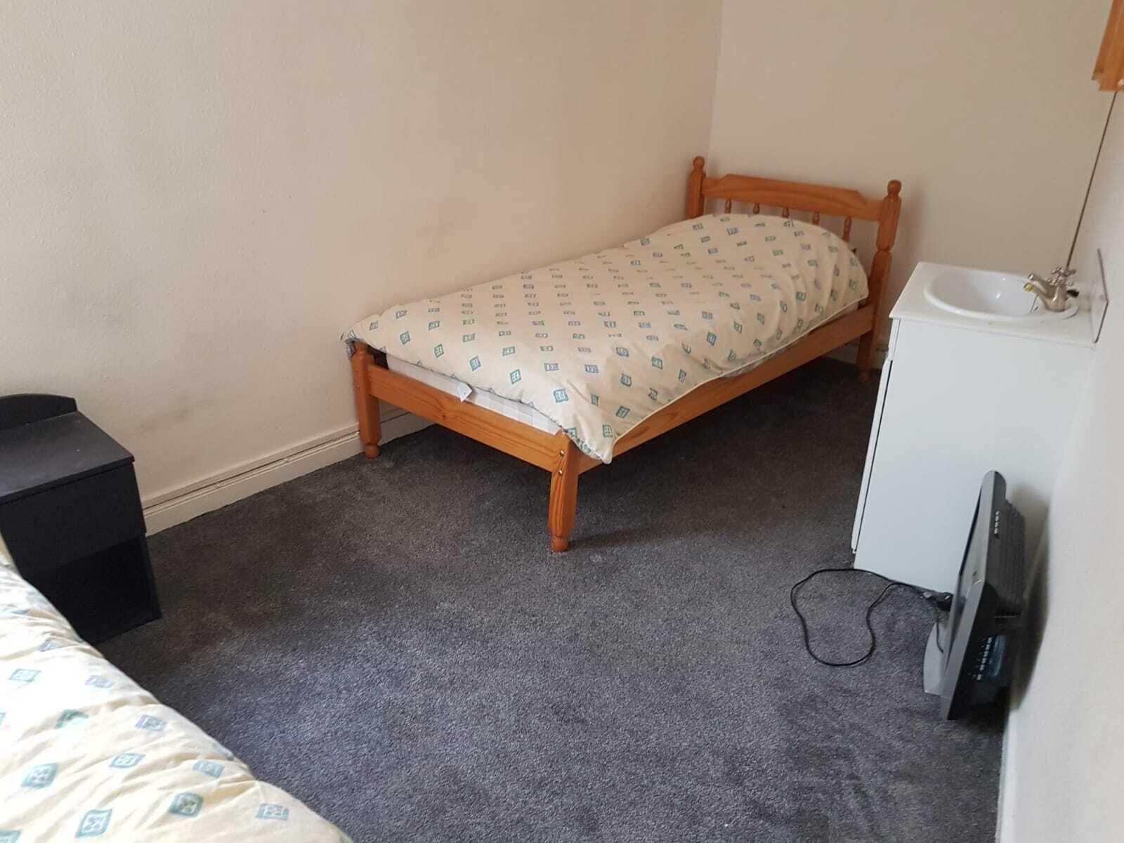 Una habitación pequeña con una cama individual, un cabecero de madera y una colcha estampada. Hay un lavabo blanco y un televisor sobre un mueble.