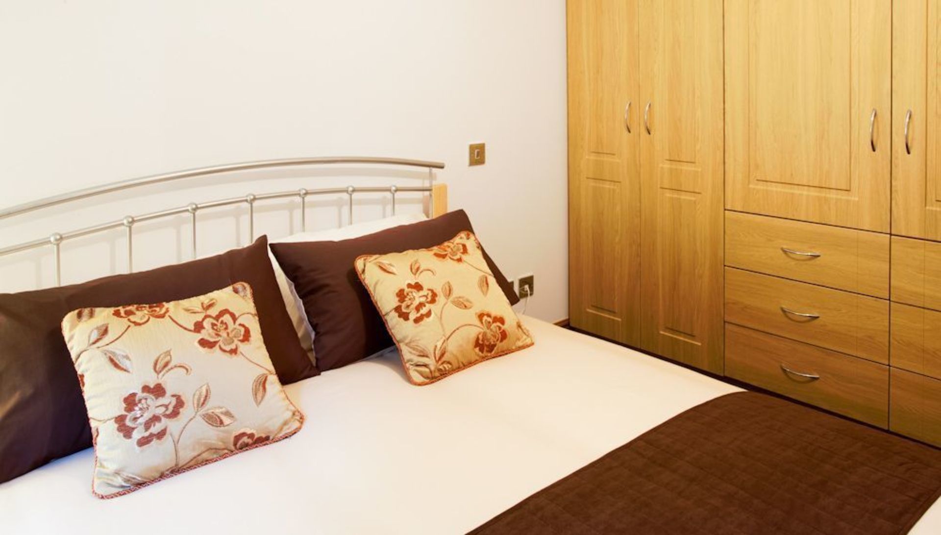 Une chambre à coucher avec un lit en métal, deux oreillers à motifs floraux, une couvre-lit marron et une armoire en bois dotée de nombreux tiroirs.