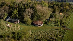 A rural landscape with a circular building surrounded by trees, a wooden structure with a sloped roof, and a fenced area with a chair.