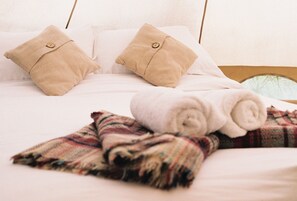 Uma cama com duas almofadas bege, uma toalha branca enrolada e um cobertor xadrez.