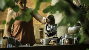 A person pouring tea from a kettle into a cup, with a rustic kitchen setting including a stove and various kitchen utensils.