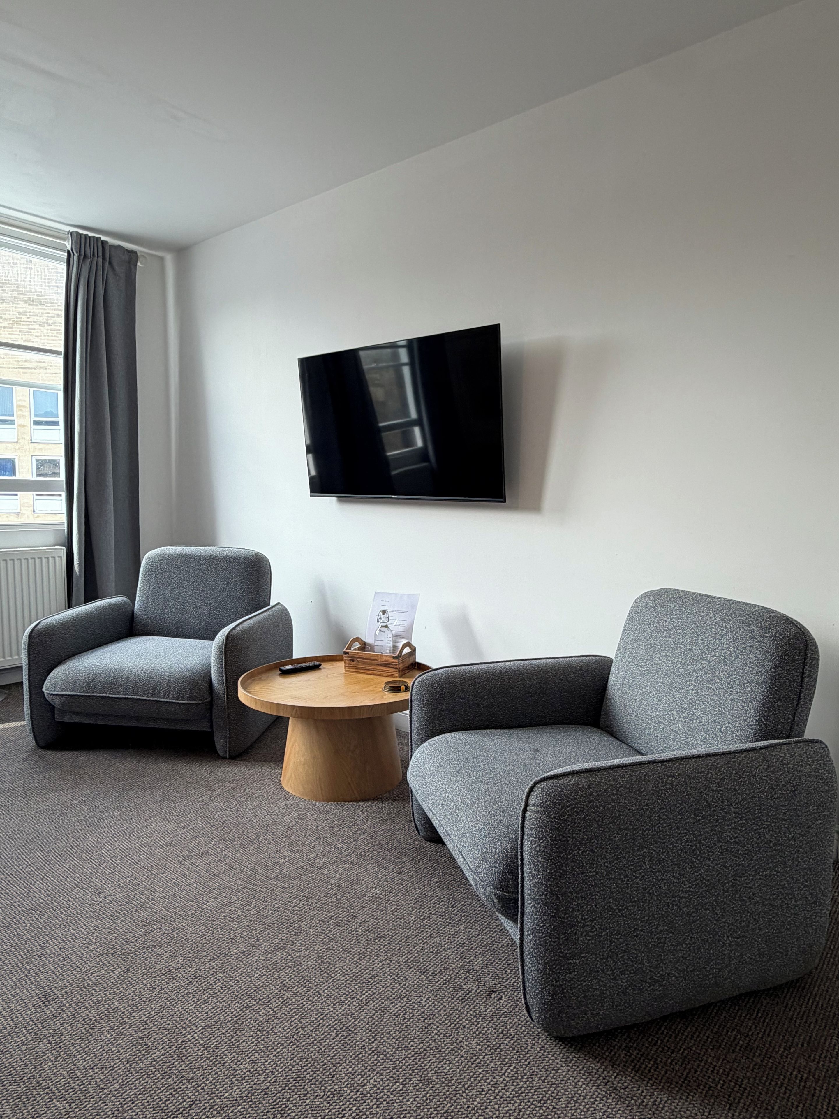 A hotel room with two grey armchairs, a wooden coffee table, a flat-screen TV mounted on the wall, and a window with curtains.