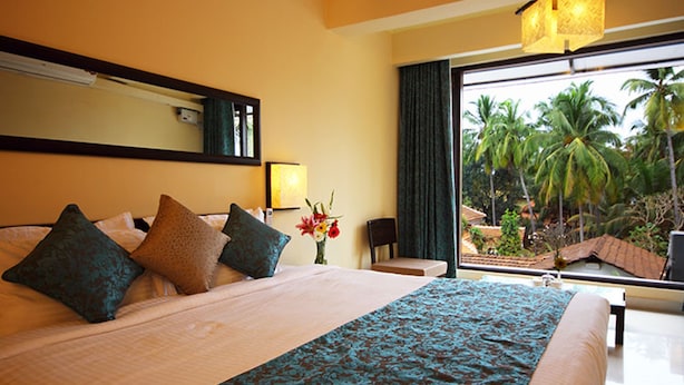 A hotel room with a large bed, a sitting area, a view of palm trees, and a window with curtains. - De Baga Deck Comforts (Calangute)