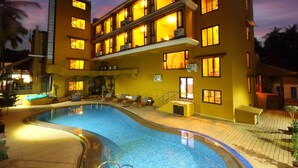 A hotel with a swimming pool, multiple balconies, and a well-lit exterior at dusk. - De Baga Deck Comforts (Calangute)