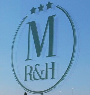 A logo with a large capital 'M' and the letters 'R&H' below it, encircled by a ring with three stars above. - Mansio Residence Hotel (Elmas)