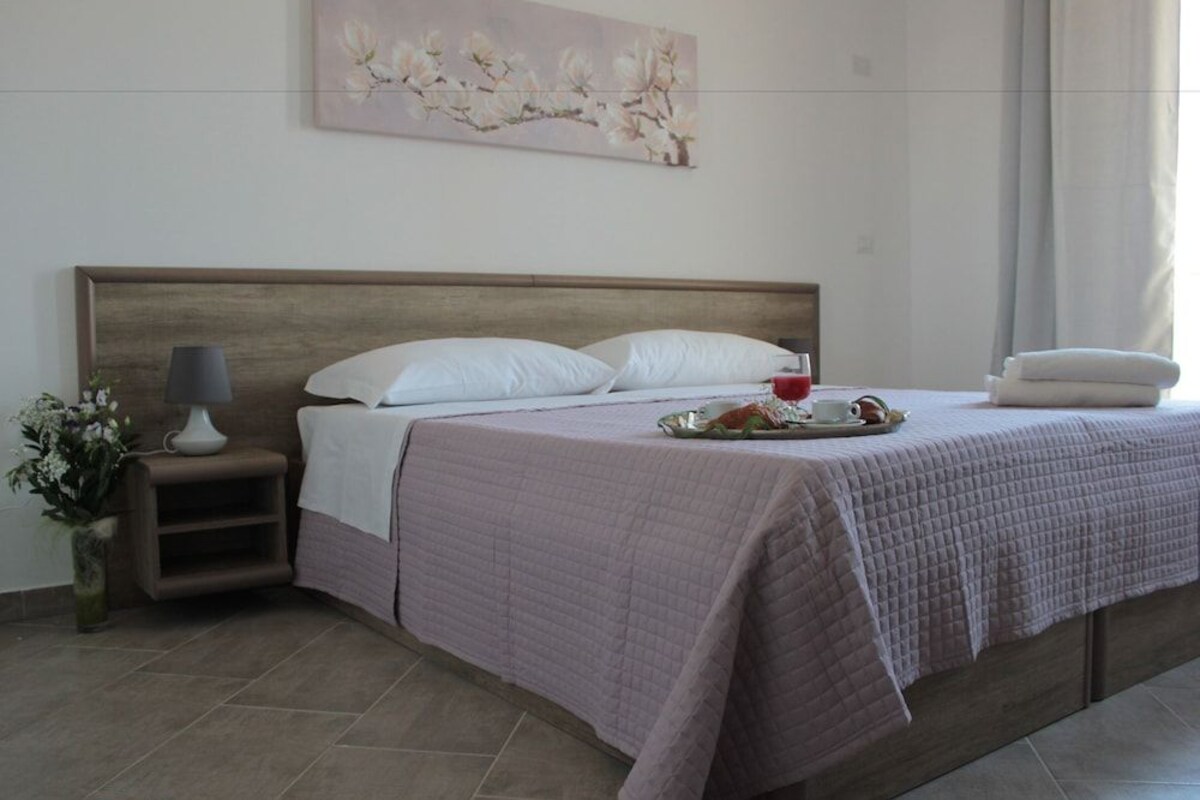 Una cama bien hecha con una colcha acolchada, una mesita de noche con una lámpara, un jarrón con flores y un cuadro en la pared.