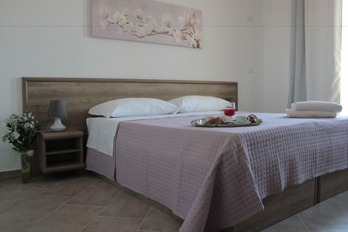 Una cama bien hecha con una colcha acolchada, una mesita de noche con una lámpara, un jarrón con flores y un cuadro en la pared.