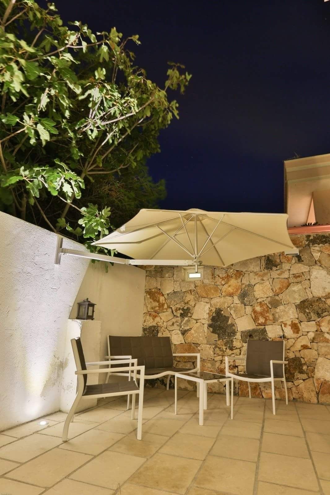 An outdoor seating area with white chairs and a large umbrella, set against a stone wall and a tree.