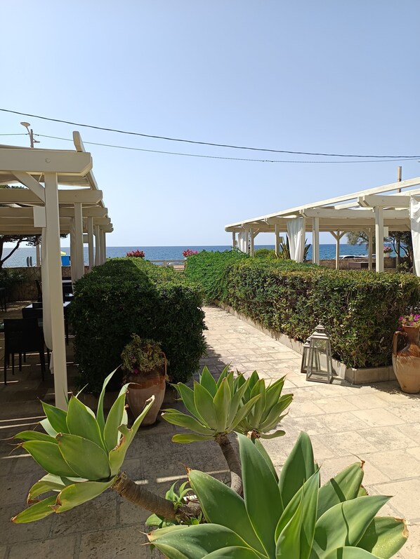 A covered outdoor seating area with a view of the sea, surrounded by greenery and potted plants.
