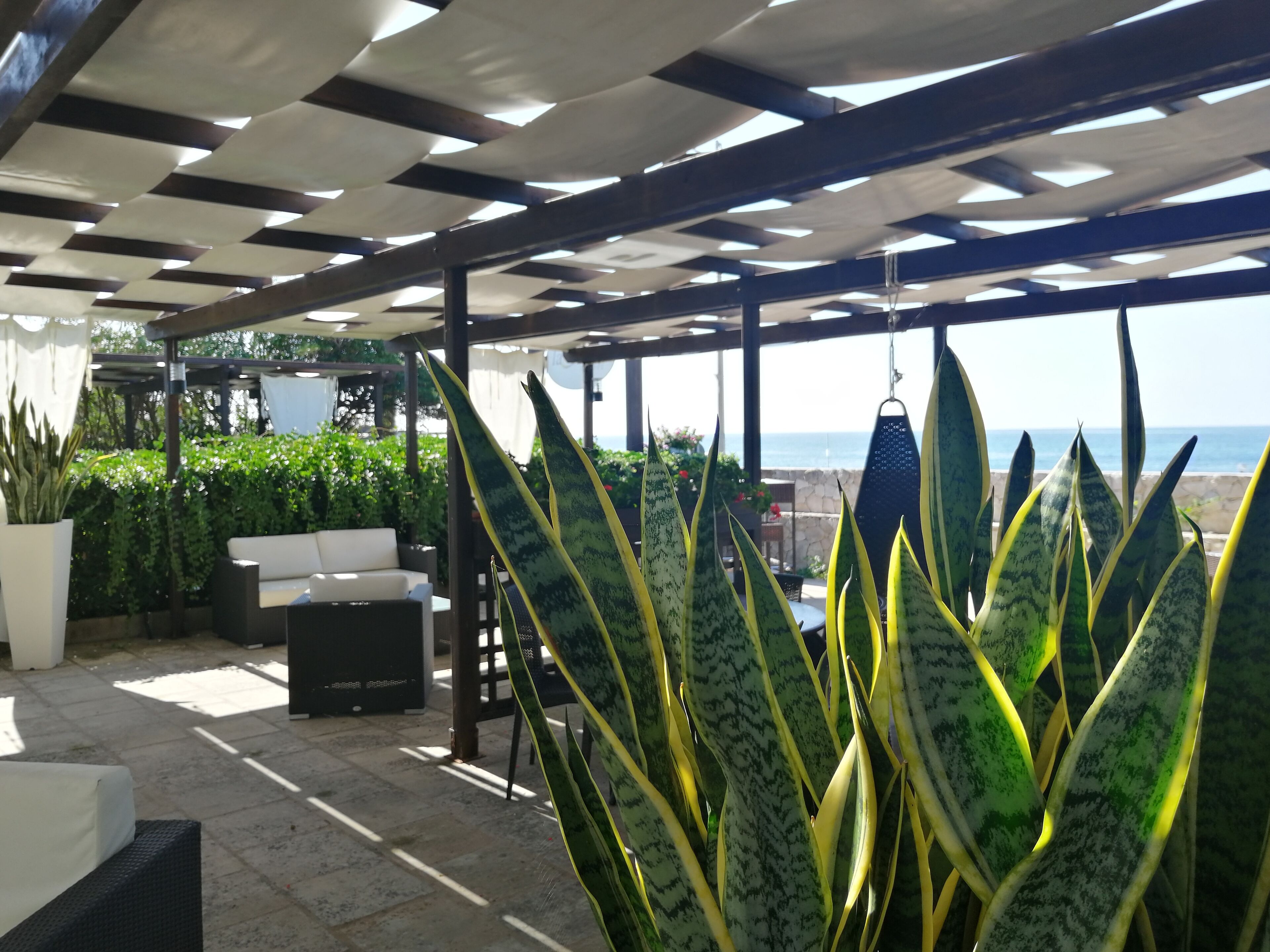 A covered outdoor seating area with a view of the ocean, featuring a pergola, potted plants, and outdoor furniture.